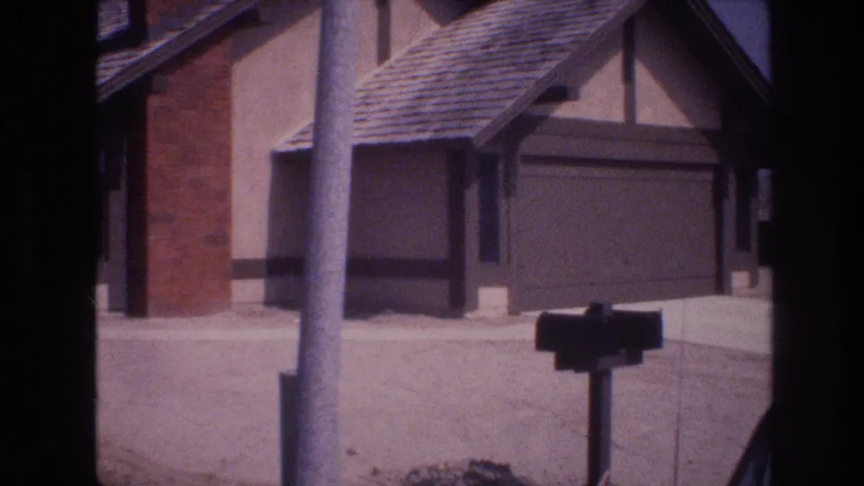 1977 Las Vegas 8mm still of a suburban home exterior, with a utility pole crossing the frame, a dark mailbox, brick chimney, and attached garage.