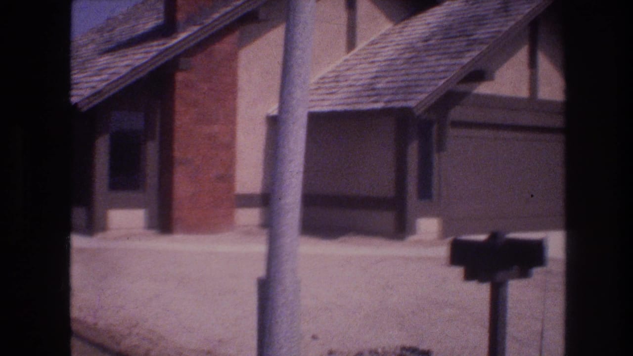 Soft 8mm frame from 1977 Las Vegas showing a brown stucco house with brick chimney, attached garage, utility pole, curbside mailbox, and sunlit street.