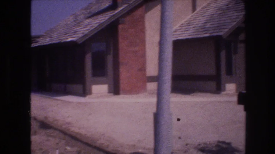 1977 Las Vegas home-movie frame showing a single-story stucco house with a brick chimney, sloped shingle roof, dry roadside ground, and a vertical pole in the foreground.