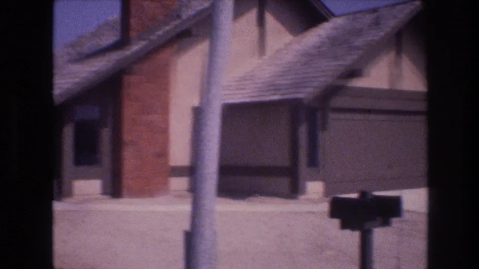 Soft 8mm frame from Las Vegas in 1977 showing a closer view of the residential facade with the brick chimney, roof shingles, small window, shaded wall, and a pole crossing the foreground.