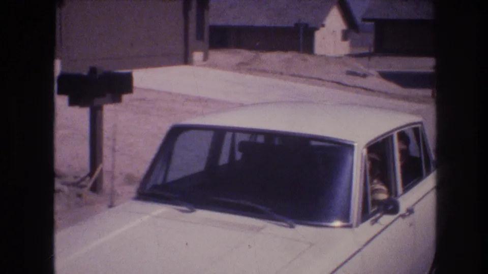 1977 Las Vegas home-movie frame showing a white sedan at the curb in front of a suburban house, with passengers visible through the windows and dry roadside ground behind it.