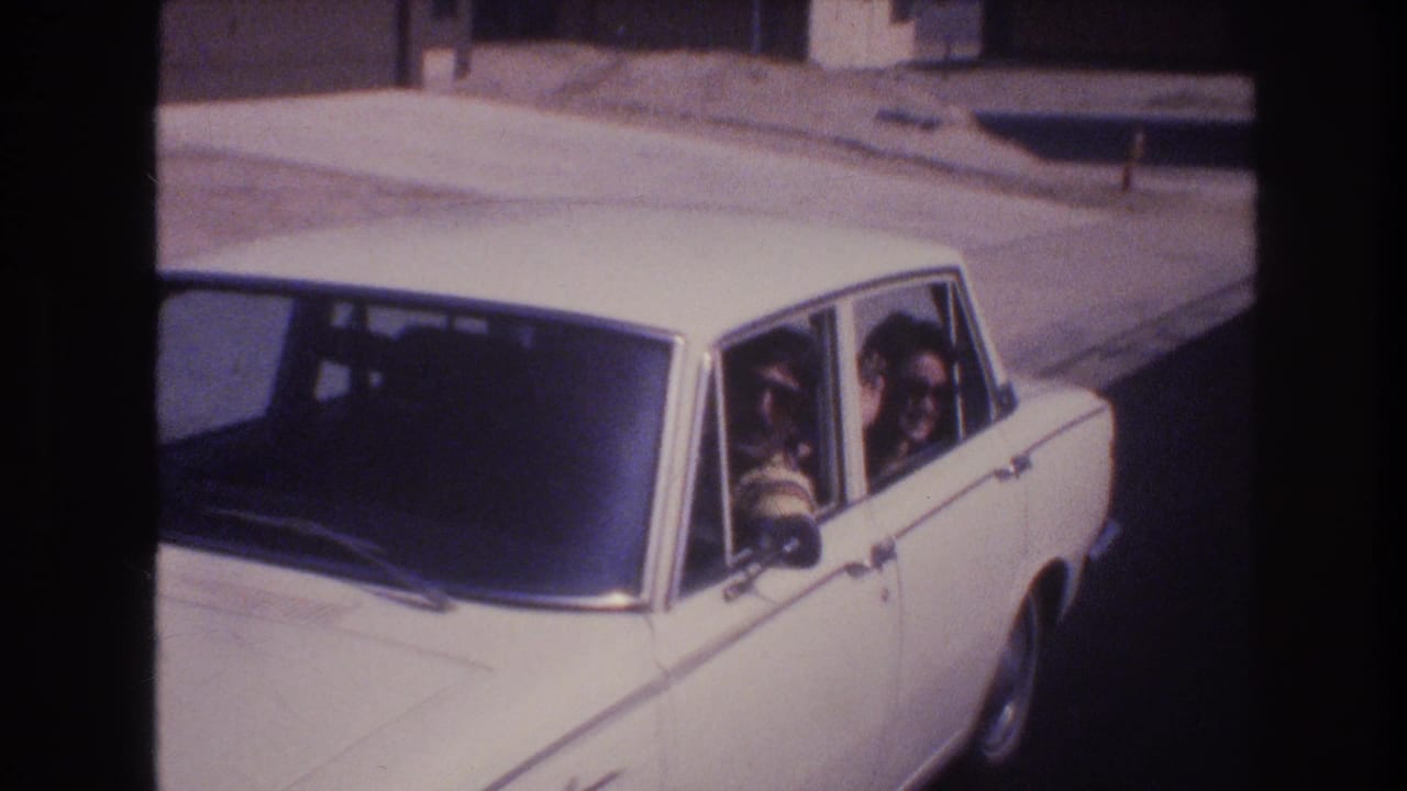A white four-door sedan drives through a Las Vegas residential street in 1977, with two adult men in sunglasses visible inside through the side windows.