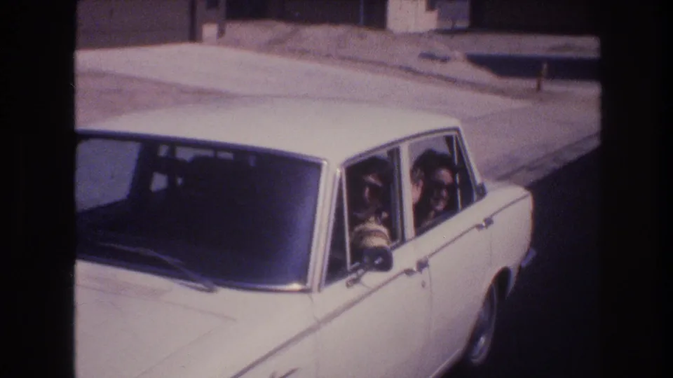 A white four-door sedan drives through a Las Vegas residential street in 1977, with two adult men in sunglasses visible inside through the side windows.