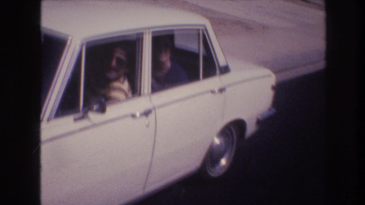 The white sedan fills more of the frame as two adult men wearing sunglasses are seen through the open side windows in the 1977 Las Vegas home-movie footage.