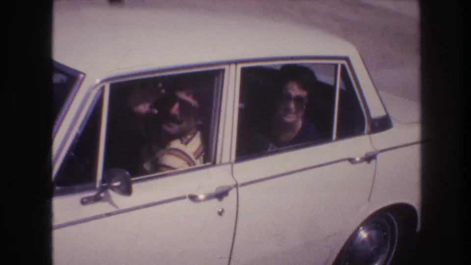 The moving white four-door sedan passes the home-movie camera, showing the driver in a striped shirt and a passenger in sunglasses against a residential street backdrop.