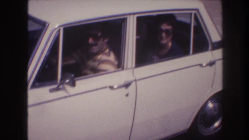 Near the end of the clip, the white sedan is close to the camera, with both adult occupants in sunglasses visible through the side windows in bright daylight.