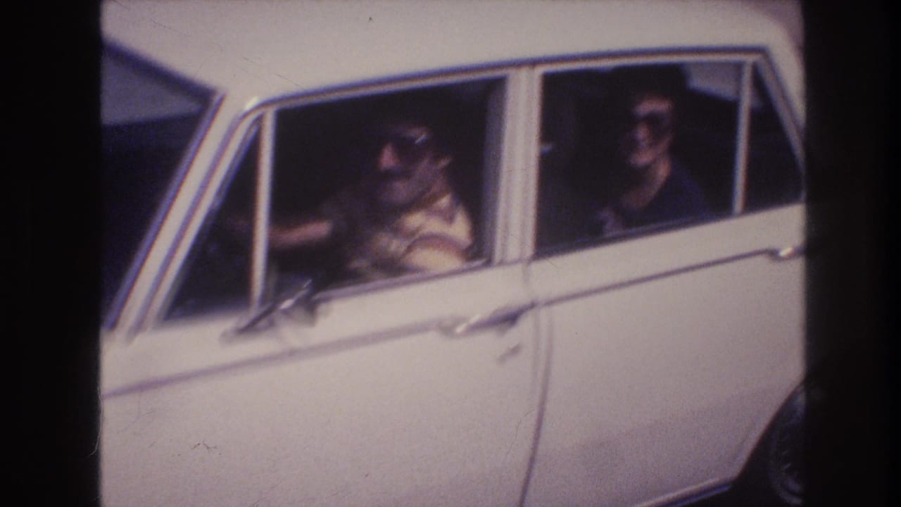 Two adults wearing sunglasses sit in a white sedan seen through the side windows.