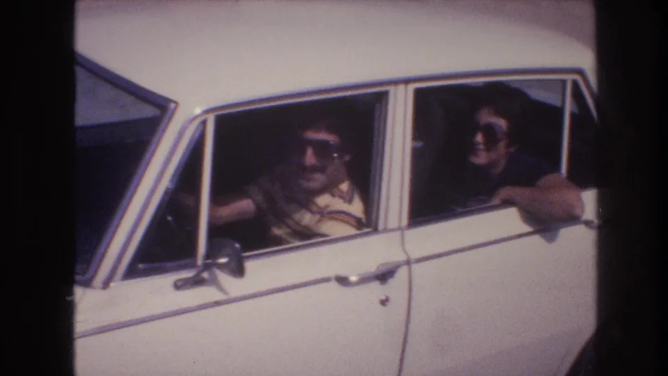 A 1970s home-movie view of two adult travelers in a white sedan, with the rear passenger leaning near the open window.
