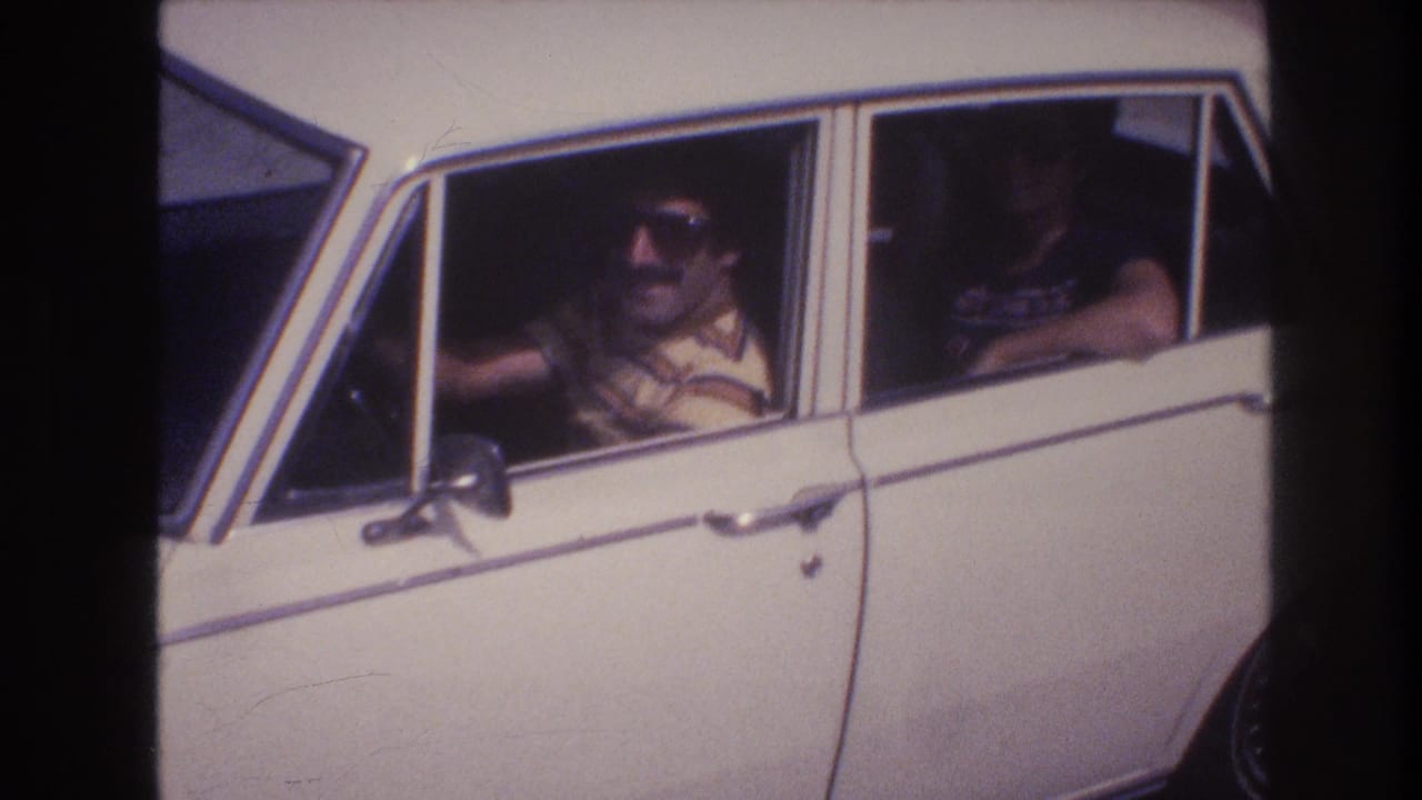 Close home-movie frame of the driver in sunglasses at the wheel of a white sedan, with a rear-seat passenger partly visible.