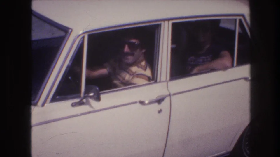 Close home-movie frame of the driver in sunglasses at the wheel of a white sedan, with a rear-seat passenger partly visible.