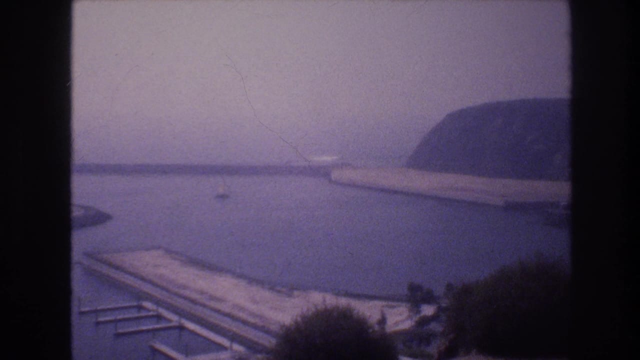 1977 8mm home-movie frame of a misty harbor scene with calm water, a long pier, a small boat, and a dark cliff or hillside in the background.