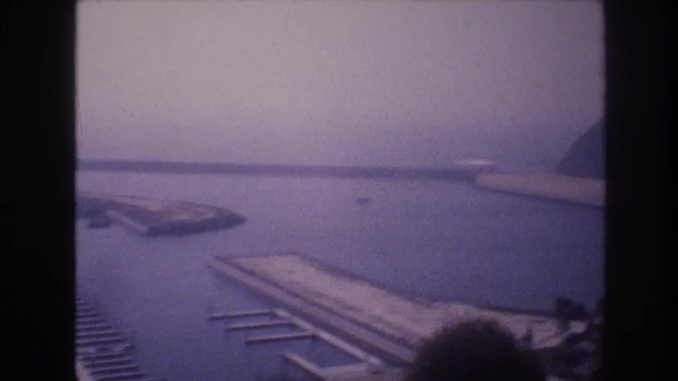 1977 8mm home-movie frame showing a soft-focus pan across harbor water, dock slips, a concrete pier, and a hazy hillside at the edge of the frame.