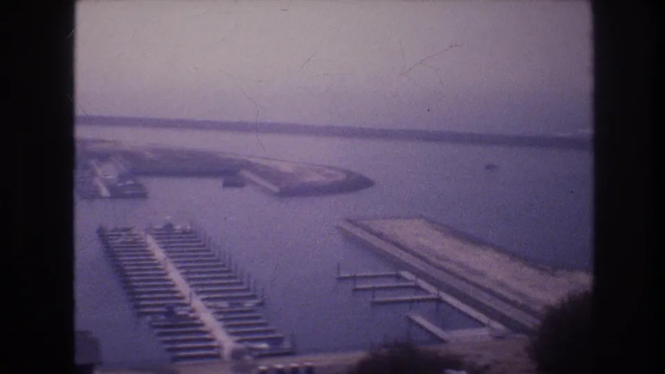 1977 8mm home-movie frame with rows of marina slips, long dock structures, calm harbor water, and a hazy shoreline in the background.