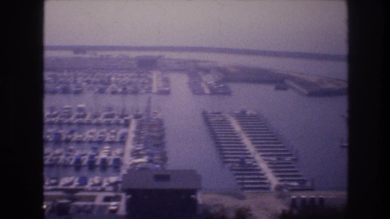 1977 8mm aerial frame of a Las Vegas-area marina with clustered sailboats, parallel docks, water channels, parking areas, and low marina structures.