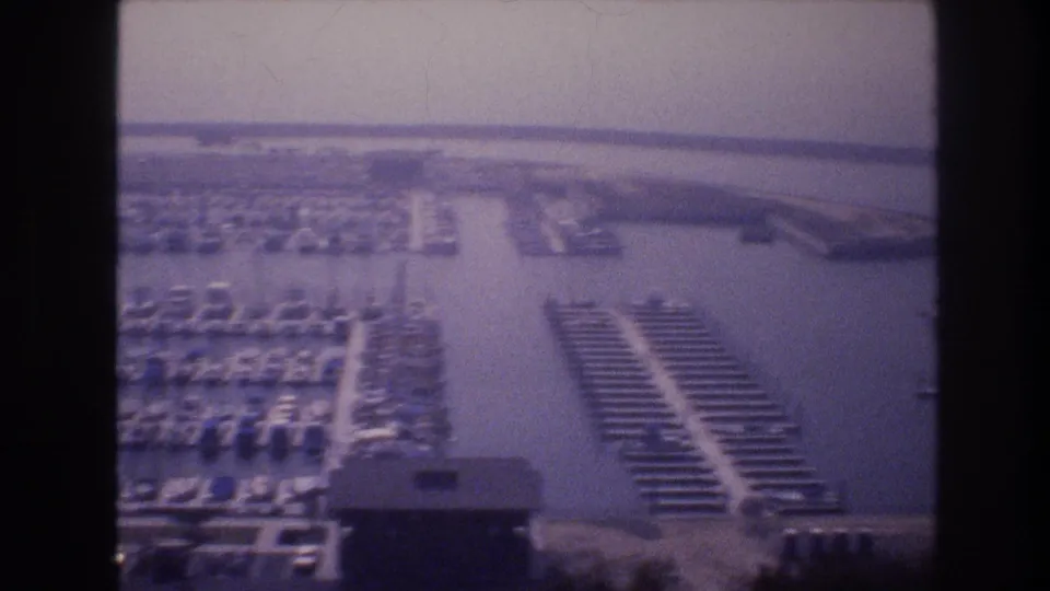 1977 8mm aerial frame of a Las Vegas-area marina with clustered sailboats, parallel docks, water channels, parking areas, and low marina structures.