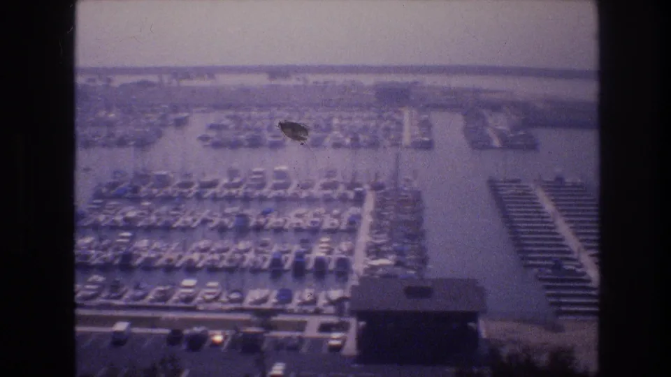 Archival 8mm aerial view of the Las Vegas-area marina, with crowded dock rows, moored boats, water lanes, shoreline roads, and a visible film blemish.