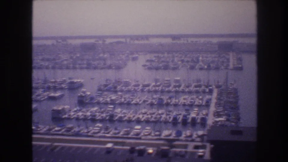 1977 Las Vegas-area aerial marina frame showing many docked boats, intersecting piers, calm water, parking areas, and low structures under a hazy sky.