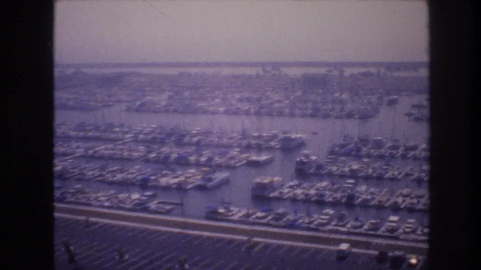 Wide 1977 Las Vegas-area marina view with clustered boats, organized docks, parking rows, and a distant shoreline visible through soft, purple-tinted film.