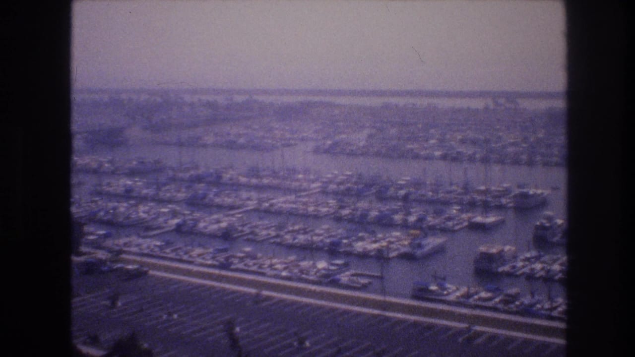 Aerial 8mm frame from 1977 showing a Las Vegas marina with crowded docks, many small boats, a broad parking area, and low land in the distance.
