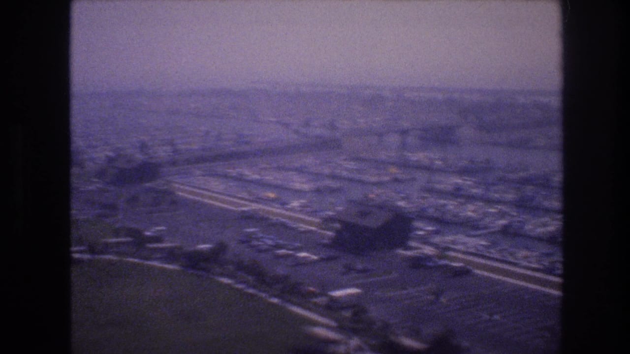 1977 Las Vegas-area aerial frame with broad highway lanes, large paved parking fields, low buildings, and hazy urban development beyond.