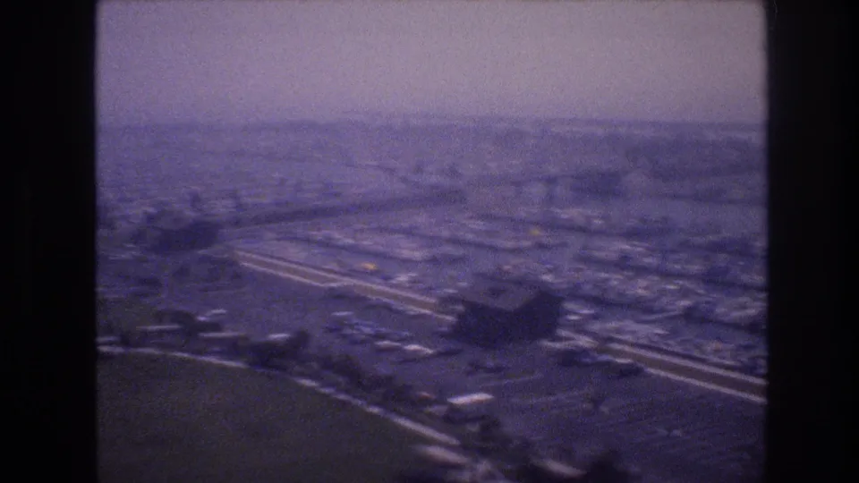 1977 Las Vegas-area aerial frame with broad highway lanes, large paved parking fields, low buildings, and hazy urban development beyond.