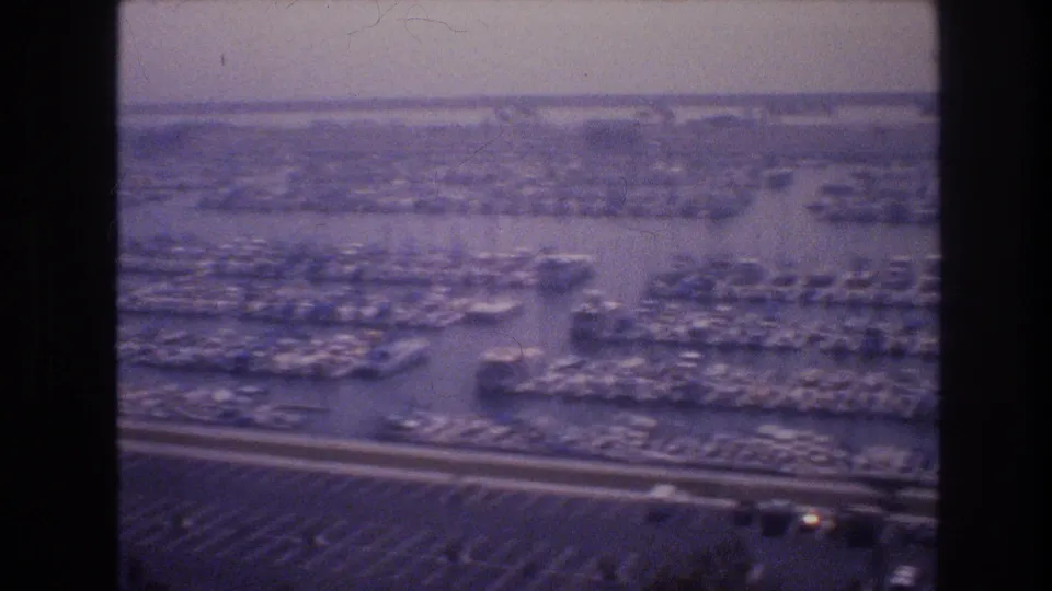 1977 Las Vegas-area aerial frame showing marina slips, numerous docked boats, adjacent roadways, parking areas, and hazy water beyond.