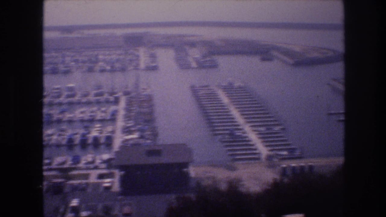 Panned aerial-perspective view of a 1970s marina showing organized boat slips, a central waterway, and a marina building, on aging 8mm film, USA, circa 1977.