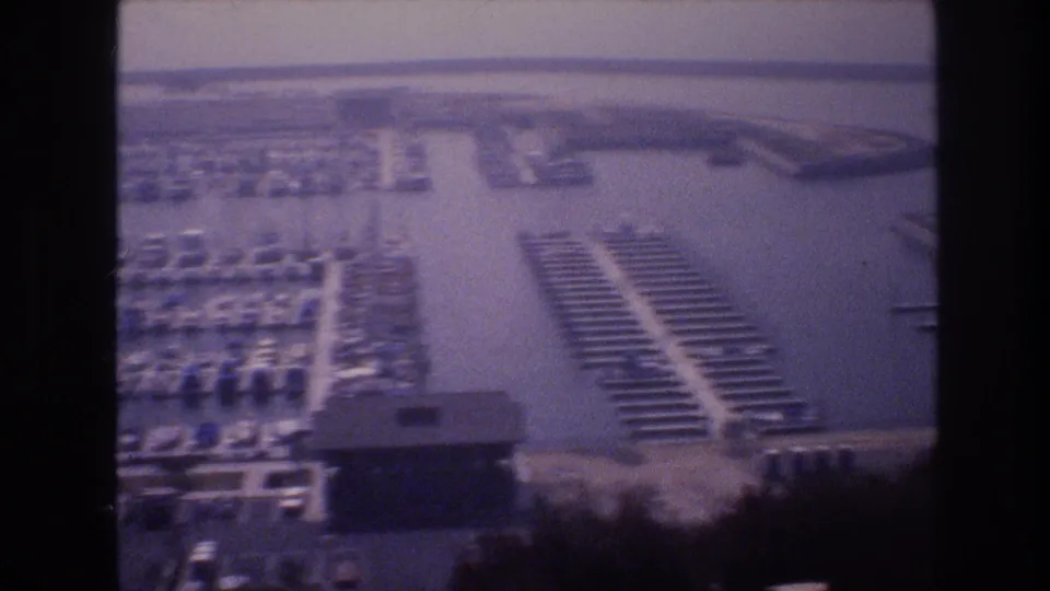 Panned aerial-perspective view of a 1970s marina showing organized boat slips, a central waterway, and a marina building, on aging 8mm film, USA, circa 1977.