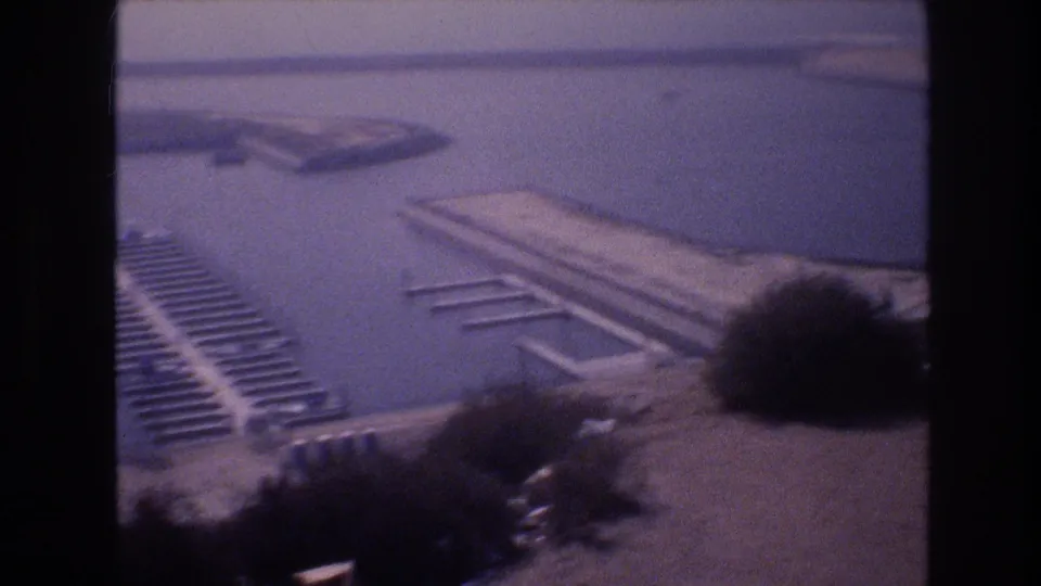 Elevated view of dock fingers mostly empty of boats, open flat water beyond, and shoreline vegetation at frame edge, faded 8mm film, USA, circa 1977.
