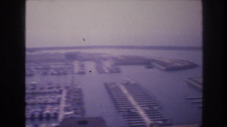 Aerial-perspective view of marina dock infrastructure and moored boats with expansive open water and low flat horizon beyond, 8mm film, USA, circa 1977.