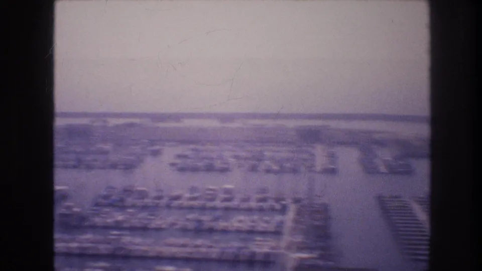 1977 8mm footage frame showing a hazy elevated view of Las Vegas with parking areas, low buildings, and distant development.