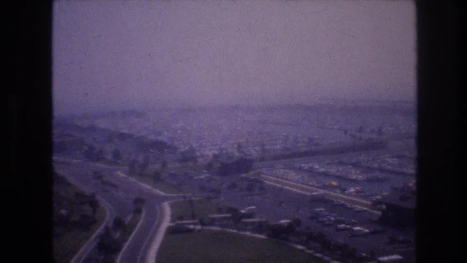 1977 8mm Las Vegas frame with curving roads, parking areas, low buildings, and a hazy city spread beyond the foreground.