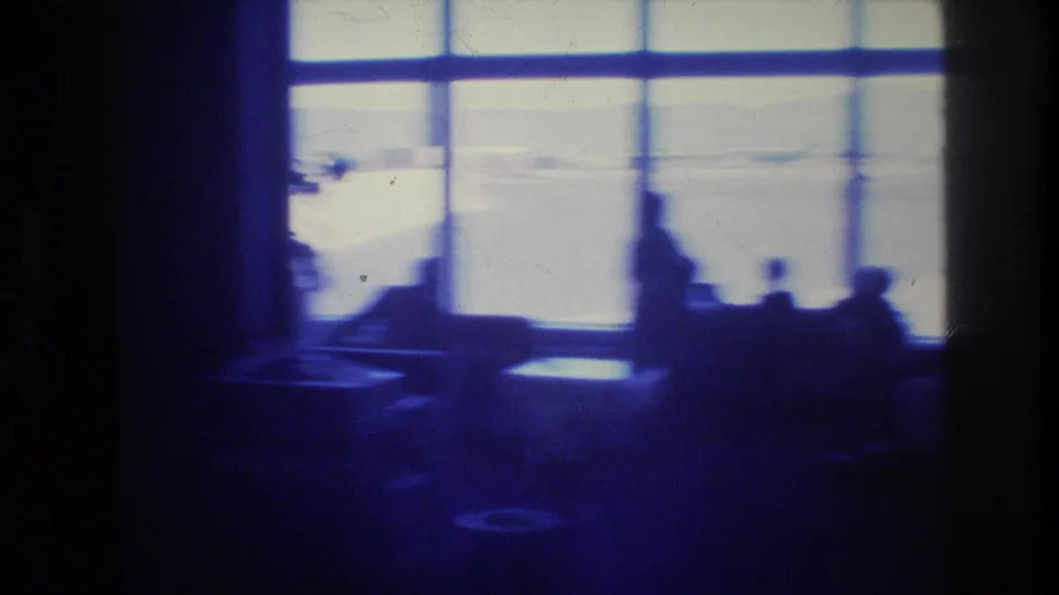 1977 Las Vegas airport terminal interior seen in silhouette, with bright windows, seating areas, and indistinct travelers in a dim blue 8mm frame.