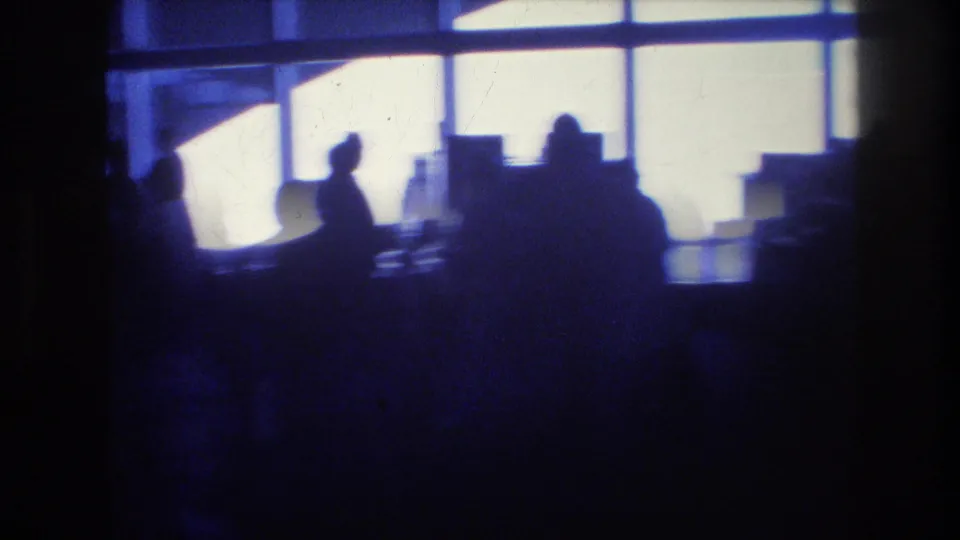 Backlit Las Vegas airport terminal scene from 1977, showing seated and standing silhouettes against large windows in a soft blue 8mm exposure.