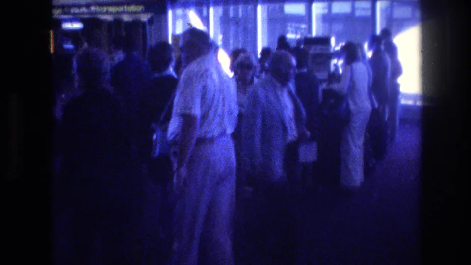 Travelers move through a dim 1977 Las Vegas airport terminal, with bright window light, overhead signage, and period clothing visible in the crowd.