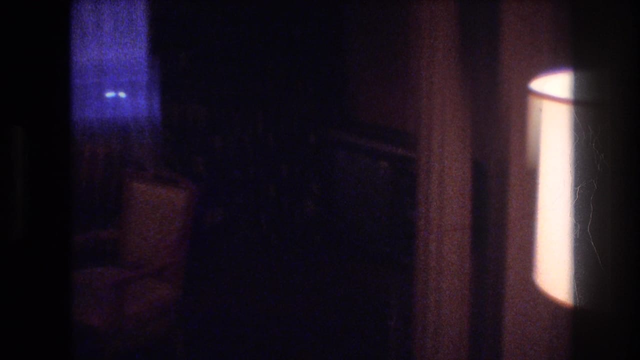 Very dark transitional interior with a dim upholstered chair at left and a bright lamp or wall light at right.