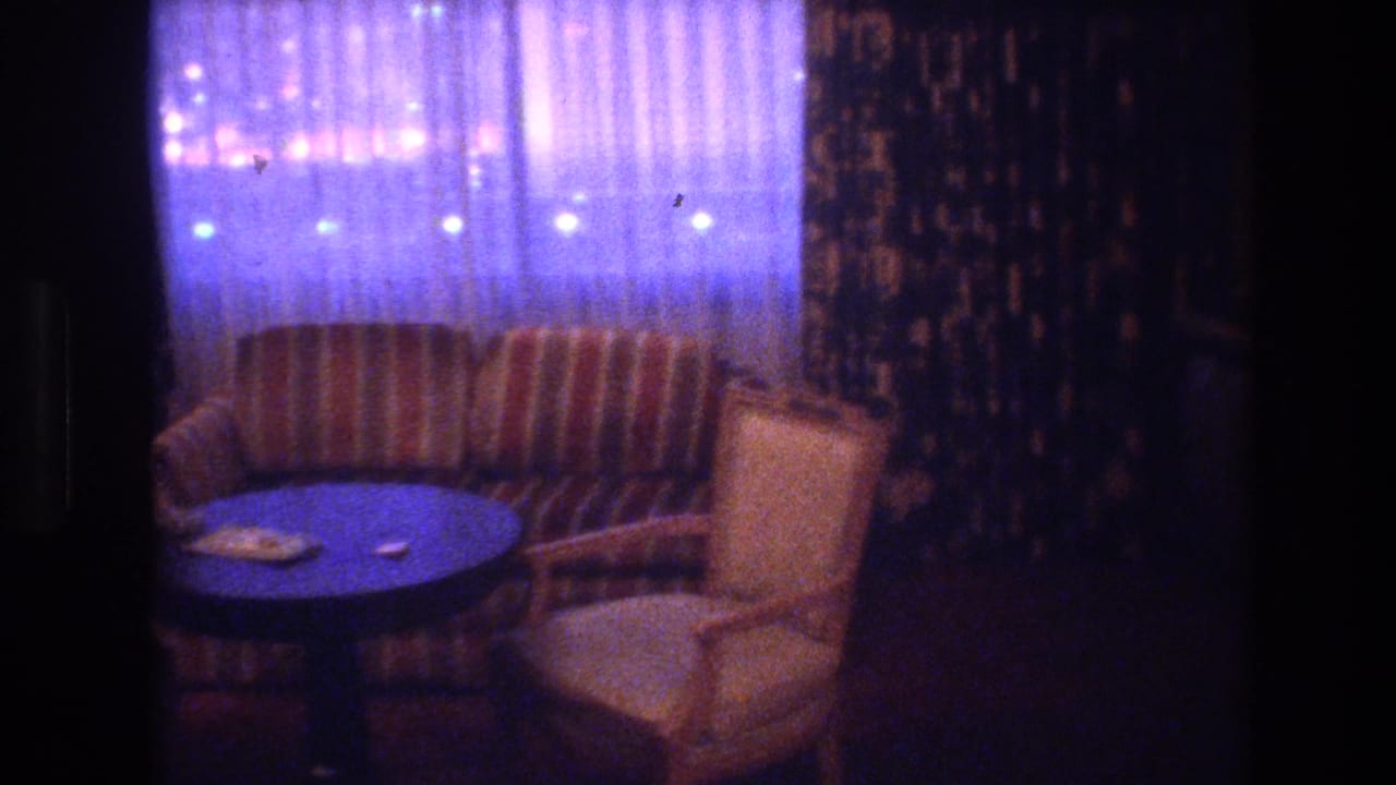 8mm hotel interior showing striped upholstered armchairs, small round table, and sheer drapes with city lights visible behind, Las Vegas, circa 1977.
