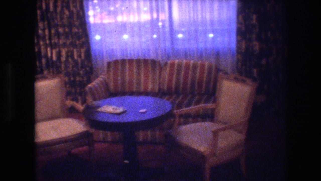 8mm interior of a 1970s Las Vegas hotel room sitting area; striped armchairs and small round table with items on the surface, sheer drapes backlit by exterior lights, circa 1977.