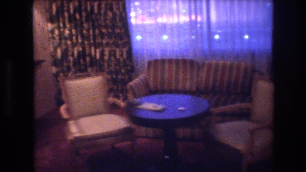 1977 8mm frame of a Las Vegas interior at night: round blue pedestal table centered, striped sofa behind, cream armchairs flanking, sheer curtains glowing blue from outside.