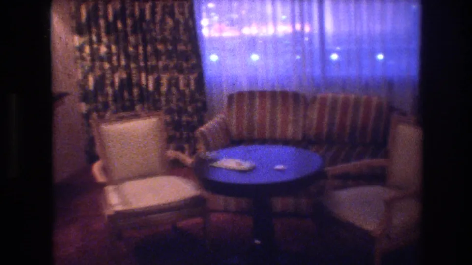 1977 8mm frame of a Las Vegas interior at night: round blue pedestal table centered, striped sofa behind, cream armchairs flanking, sheer curtains glowing blue from outside.