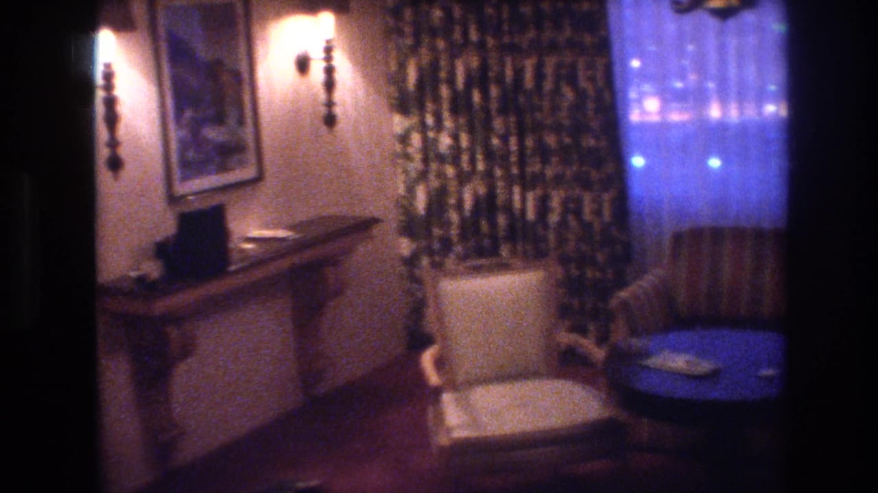 1977 8mm frame of a Las Vegas interior panned right, showing a warm-lit wall sconce, framed artwork, console sideboard, and sheer curtains glowing blue from exterior lights.