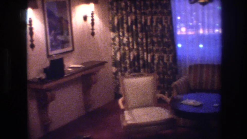 1977 8mm frame of a Las Vegas interior panned right, showing a warm-lit wall sconce, framed artwork, console sideboard, and sheer curtains glowing blue from exterior lights.
