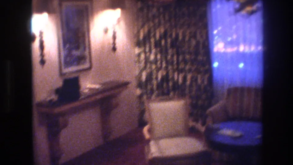 1977 8mm frame of a Las Vegas night interior: warm wall sconce and framed artwork on the left wall, console table below, sheer curtains glowing blue from exterior at right.