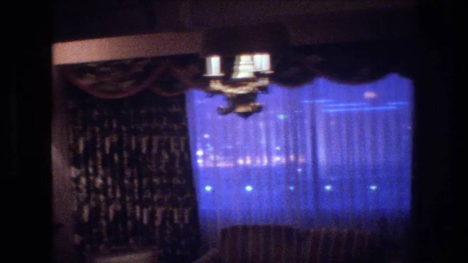 1977 8mm frame focused on sheer curtains glowing blue from exterior Las Vegas lights; a ceiling chandelier and dark patterned valance are visible above, with a wall sconce at right.