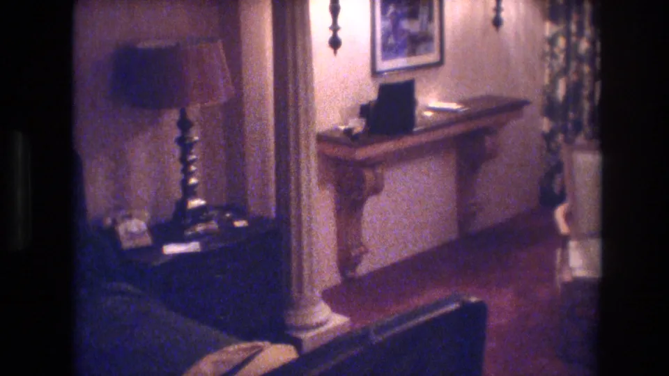 8mm film frame from 1977 Las Vegas: spindle-leg table lamp, carved console table, candlestick wall sconce, framed artwork, patterned curtains, dark blue armchair, decorative column.