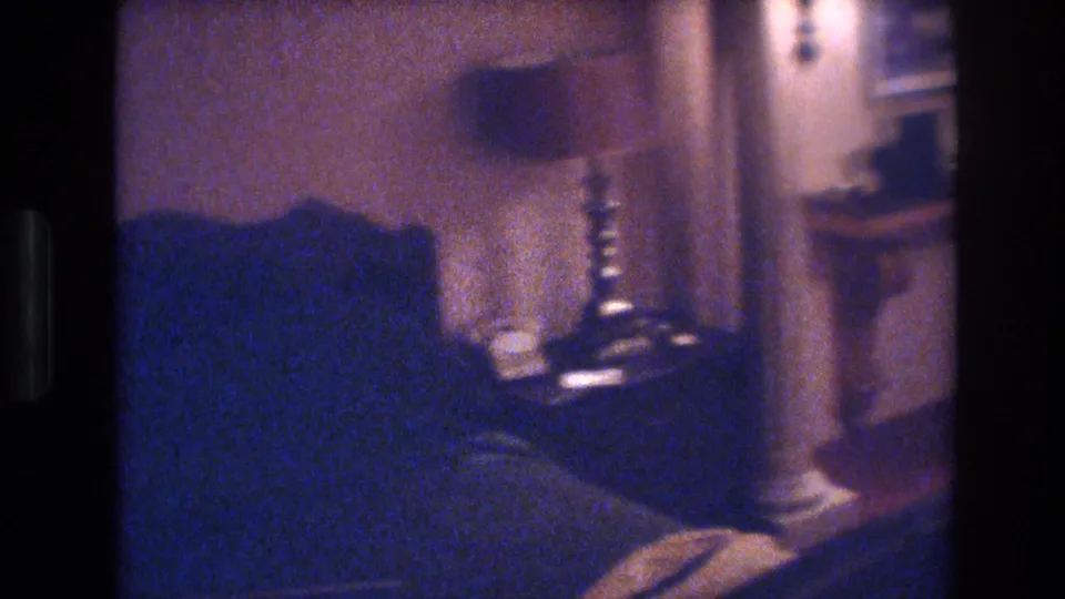 8mm film frame from 1977 showing dark blue upholstered armchair or sofa in foreground, spindle lamp base, wall sconce, and framed artwork in a warm Las Vegas room.