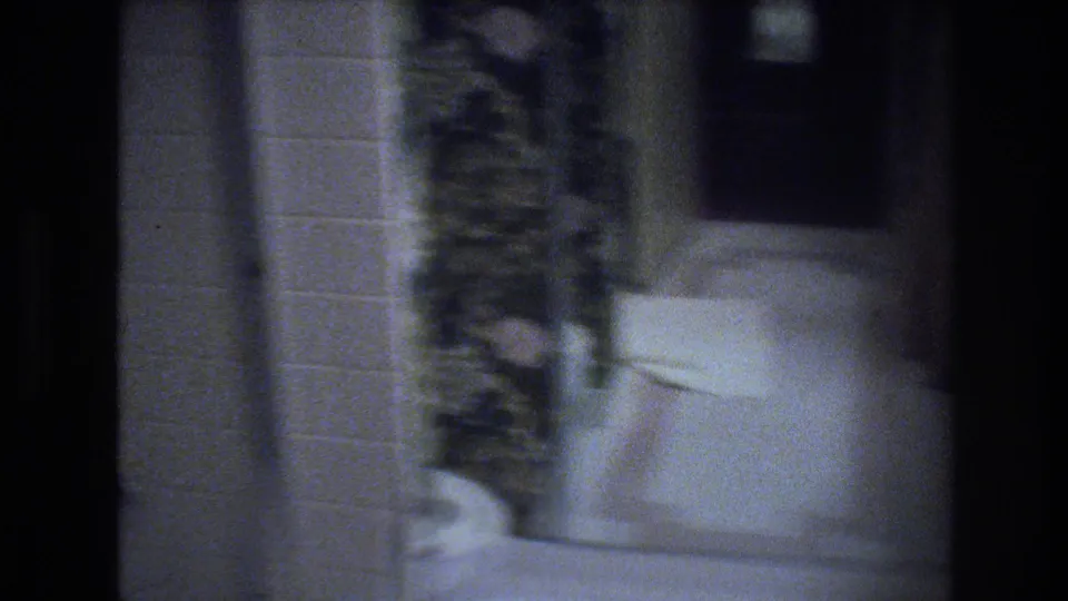 Hotel or motel bathroom, likely 1970s — white tile walls, white porcelain fixture, dark plant or foliage decor at left; camera pan produces pronounced motion blur.