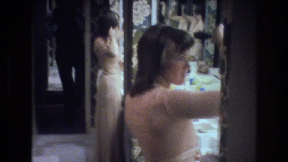 Two women in light dresses in a 1970s interior with floral decor — the background figure appears to look into a mirror in a candid home-movie frame consistent with Las Vegas travel footage.