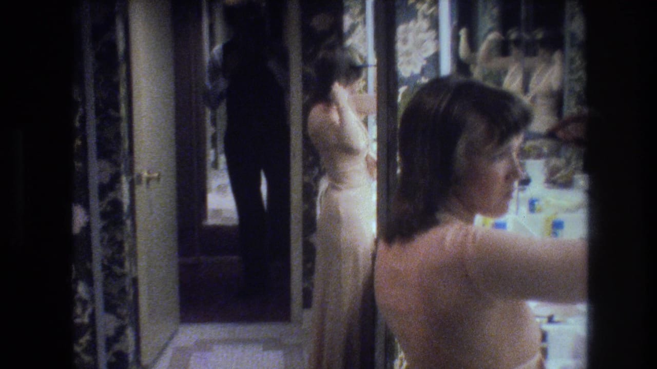 Woman with brown hair in a cream backless gown seen from behind at a mirror, with dark floral wallpaper and white tiled wall visible.