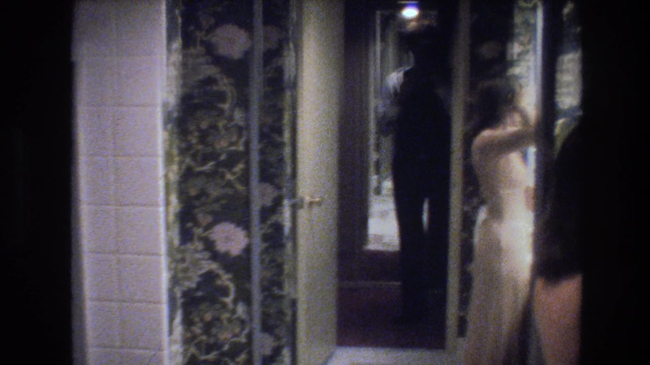 Woman in cream dress viewed from behind near a mirror; man in dark clothing visible at frame edge holding a camera; floral wallpaper and overhead light visible.